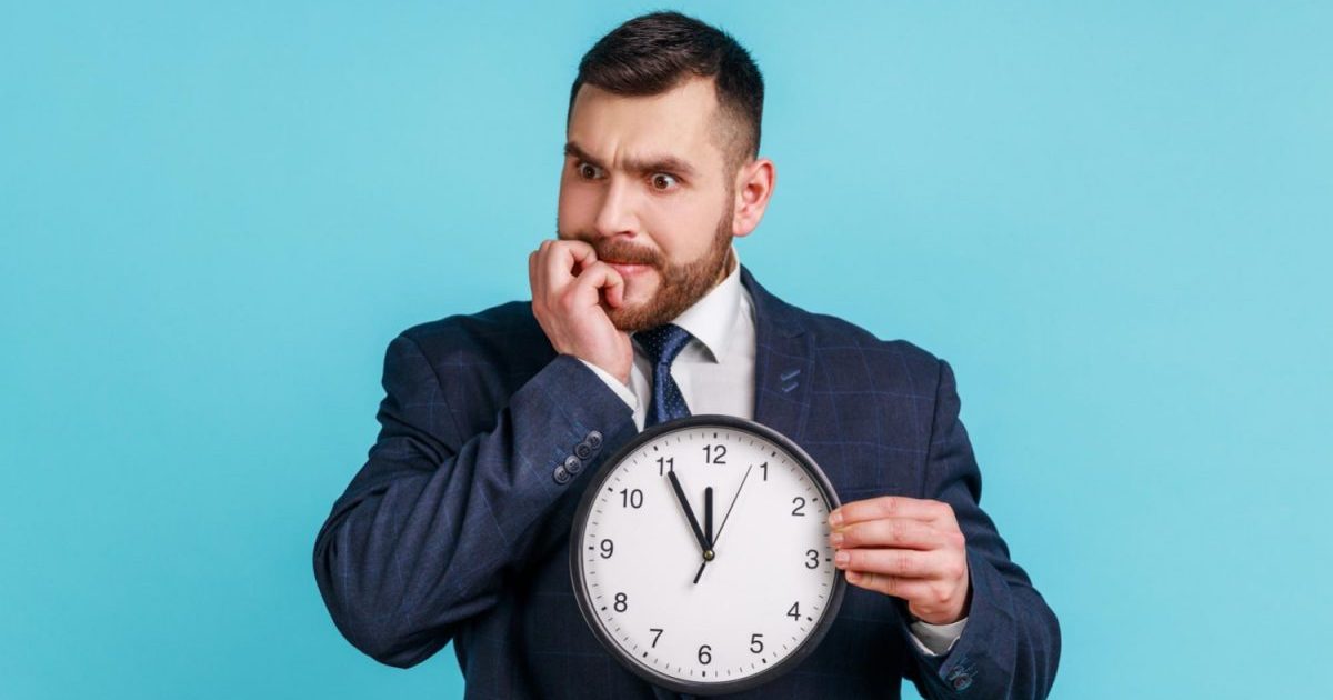 portrait-impatient-anxious-businessman-dark-suit-holding-big-clock-biting-nails-nervous-about-delayed-meeting-deadline-need-hurry-up-indoor-studio-shot-isolated-blue-background
